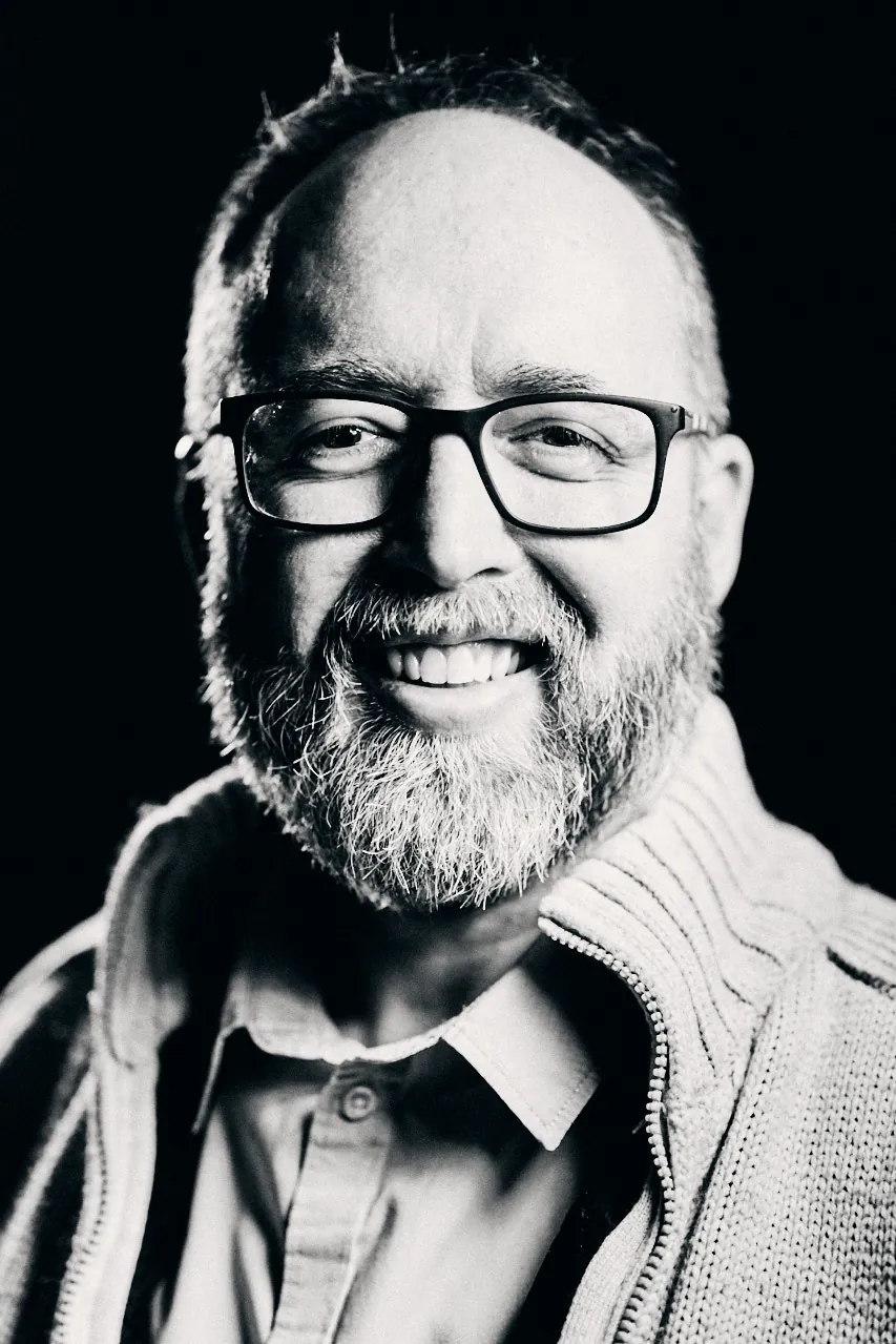 Portrait studio noir et blanc d'un homme barbu à lunettes, souriant.